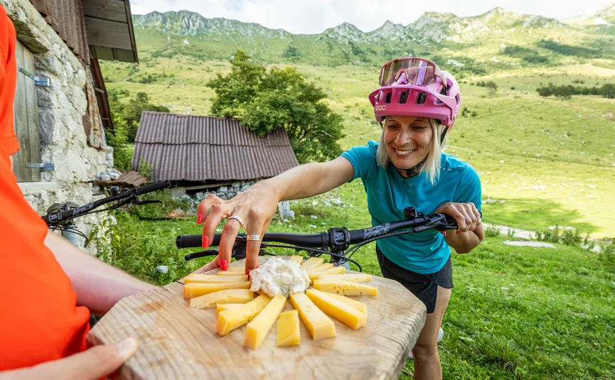 Mountainbike Urlaub in den Julischen Alpen © Soca Valley Zaprikraj - Ziga Koren Frau in Fahrradhelm greift in den Bergen nach Käse auf Holzbrett