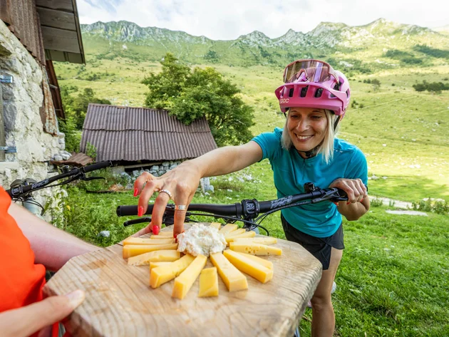 Ein perfekter Enduro-Tag – vom ersten Höhenmeter bis zum letzten Trail © Soca Valley Zaprikraj - Ziga Koren Frau in Fahrradhelm greift in den Bergen nach Käse auf Holzbrett