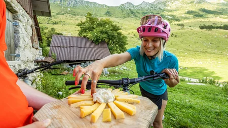 Ein perfekter Enduro-Tag – vom ersten Höhenmeter bis zum letzten Trail © Soca Valley Zaprikraj - Ziga Koren Frau in Fahrradhelm greift in den Bergen nach Käse auf Holzbrett