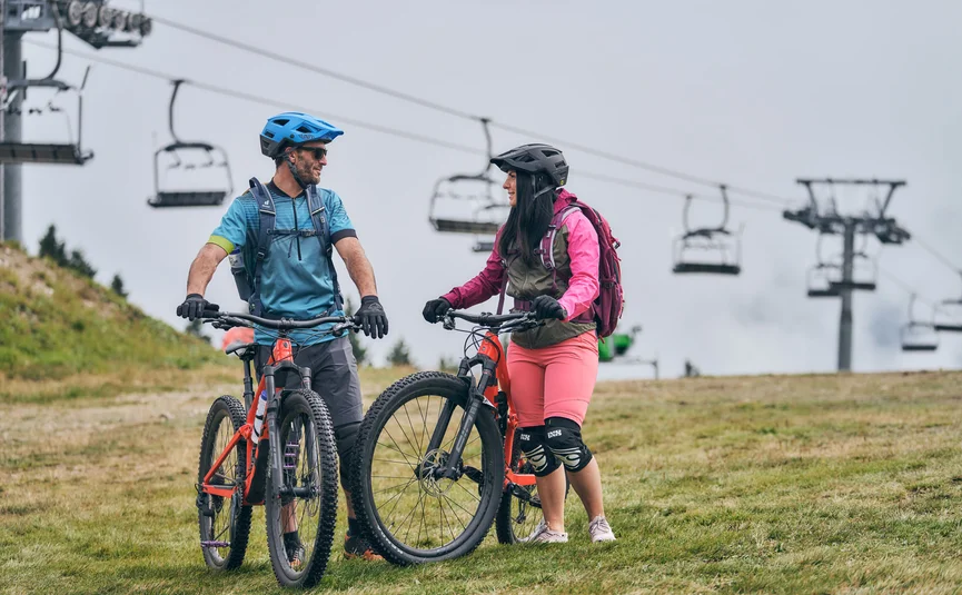 Two mountain bikers wearing helmets and gear standing with bikes on a grassy hill