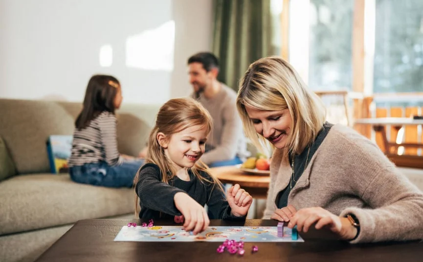 Moeder en dochter spelen bordspel, vader en kind zitten op bank op de achtergrond