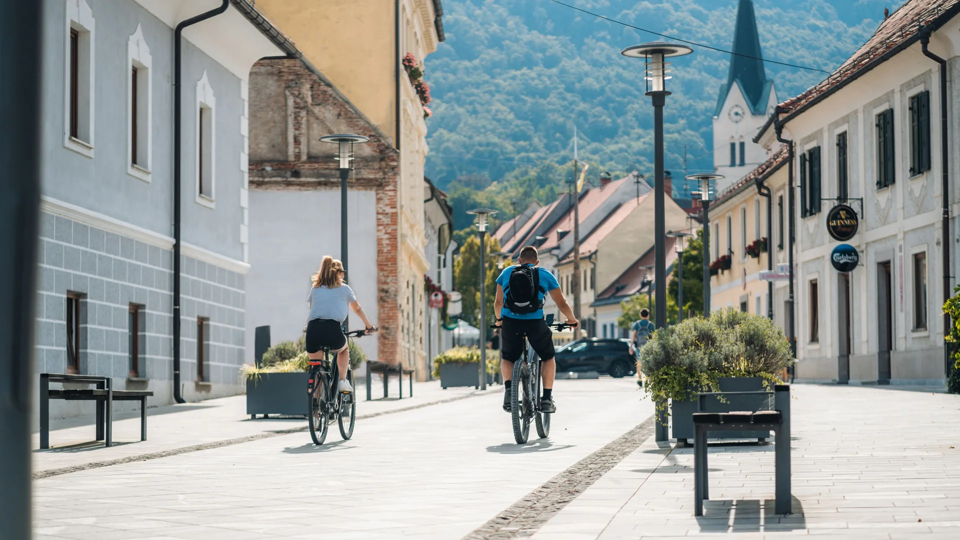 Zwei Personen fahren Fahrrad durch eine ruhige Stadtstraße mit Bergen im Hintergrund