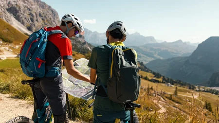 Zwei Radfahrer mit Helmen und Rucksäcken schauen auf eine Landkarte in den Bergen