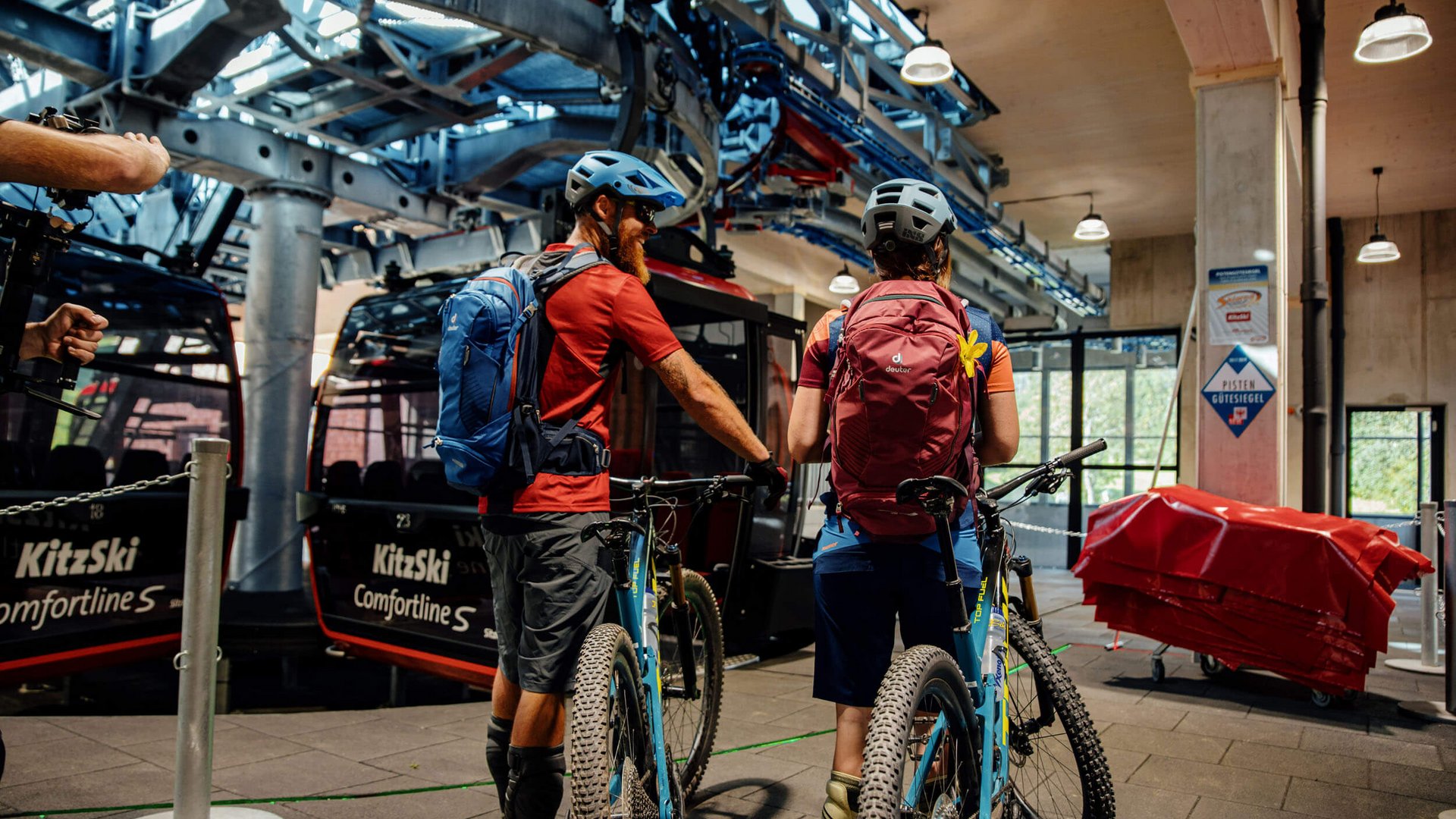 Zwei Radfahrer mit Helmen und Rucksäcken warten an der Seilbahnstation KitzSki