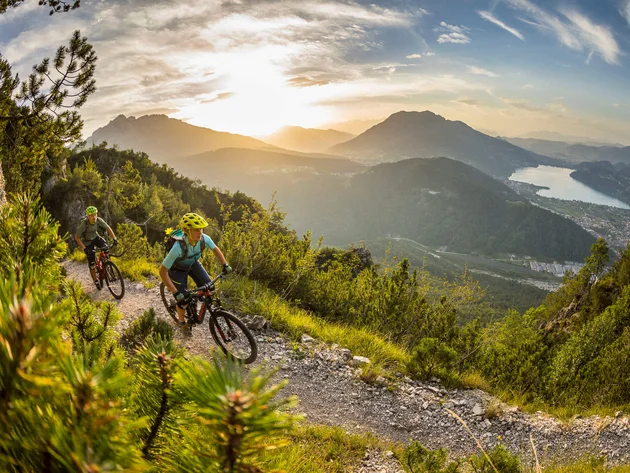Zwei Mountainbiker fahren auf einem Bergpfad bei Sonnenuntergang mit Seeblick