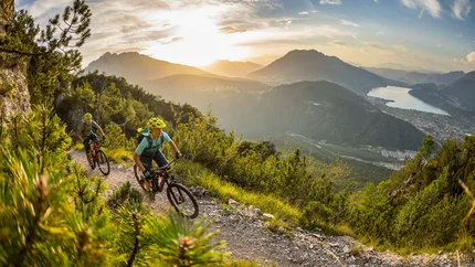 Two mountain bikers riding on a mountain trail at sunset with lake view