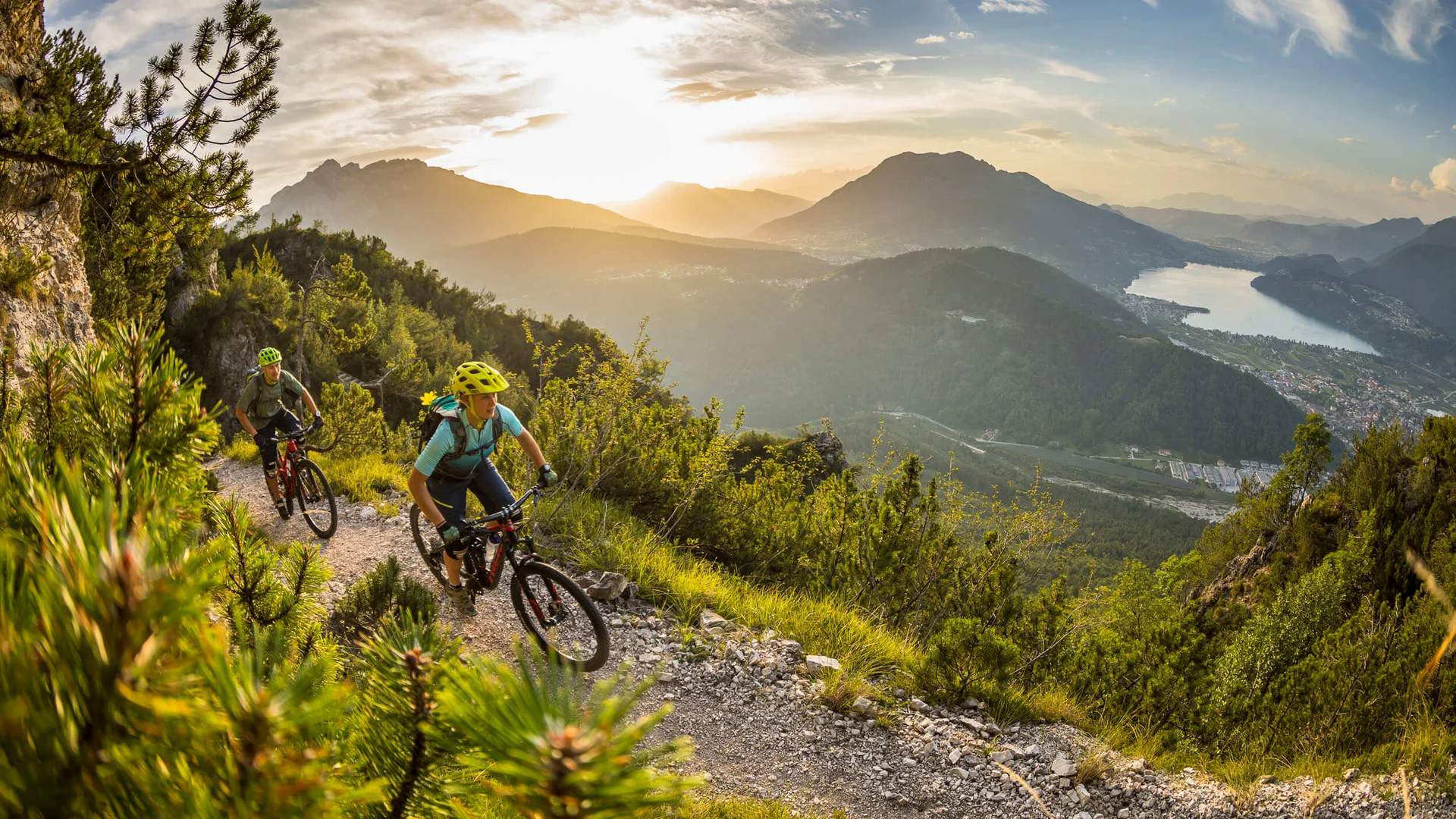 Two mountain bikers riding on a mountain trail at sunset with lake view