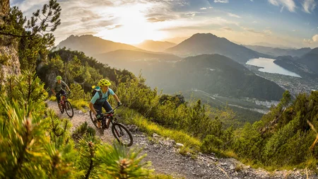 Zwei Mountainbiker fahren auf einem Bergpfad bei Sonnenuntergang mit Seeblick