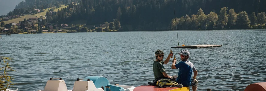 Zwei Radfahrer auf Steg am See mit Bergen im Hintergrund stoßen an