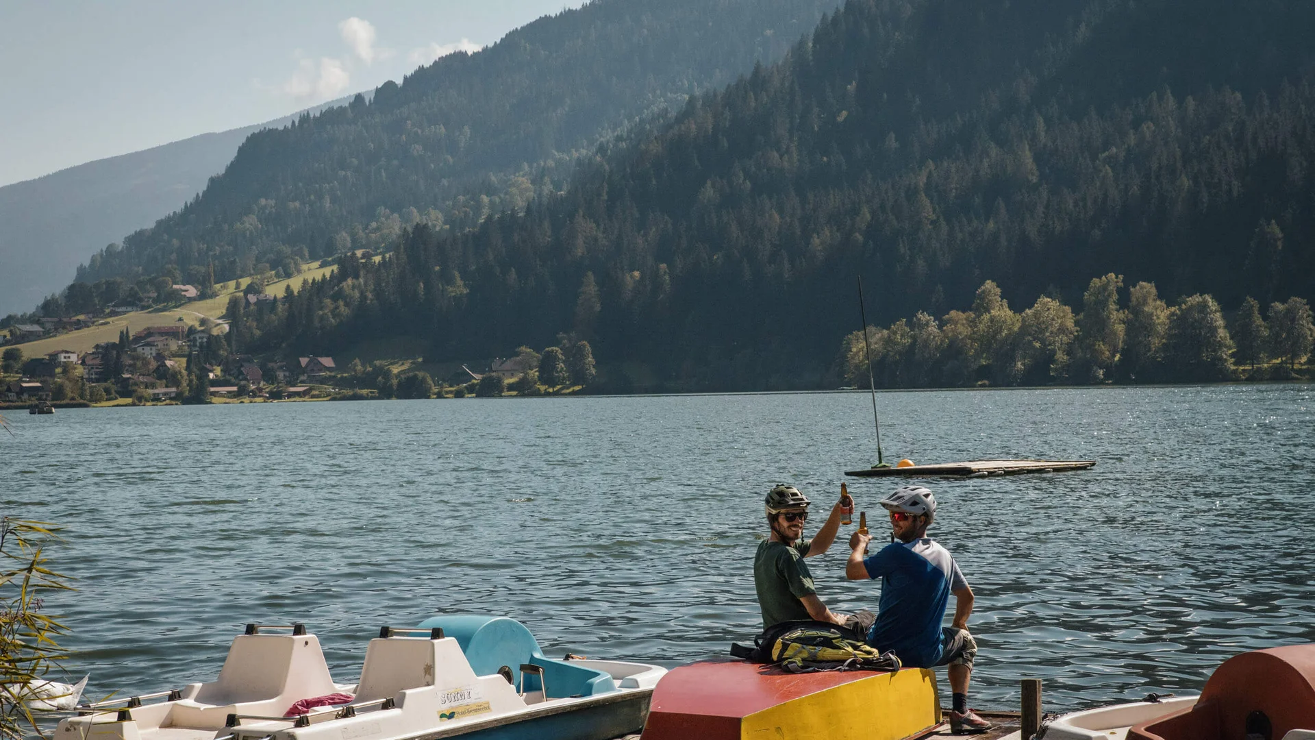 Zwei Radfahrer auf Steg am See mit Bergen im Hintergrund stoßen an