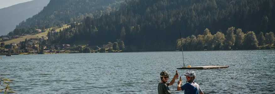 Zwei Radfahrer auf Steg am See mit Bergen im Hintergrund stoßen an