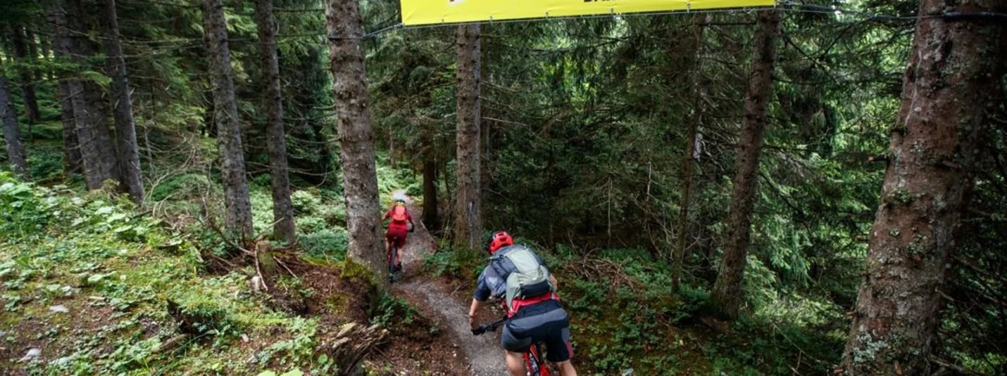 Two mountain bikers riding on a forest trail under an OurTrail banner