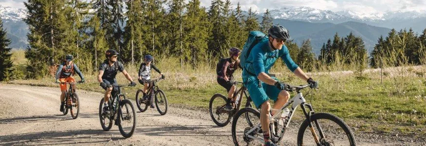 Five cyclists riding mountain bikes on a forest trail with mountains behind