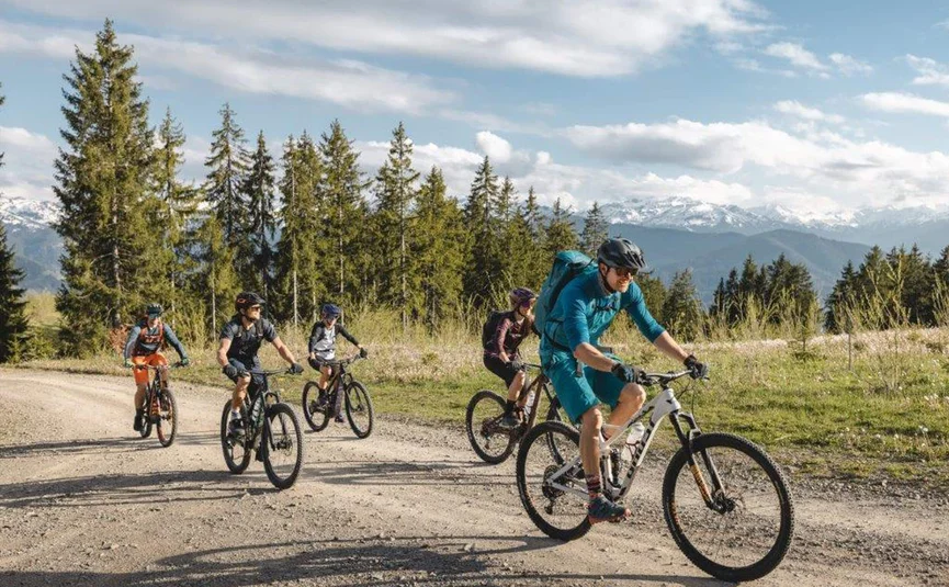 Five cyclists riding mountain bikes on a forest trail with mountains behind
