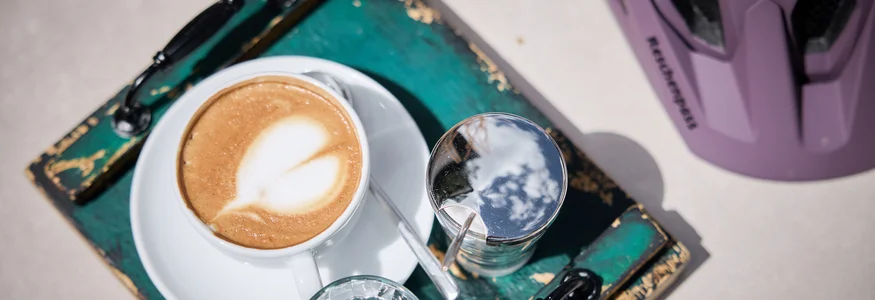 Kaffeetasse mit Milchschaum und Wasser auf einem grünen Tablett neben Fahrradhelm