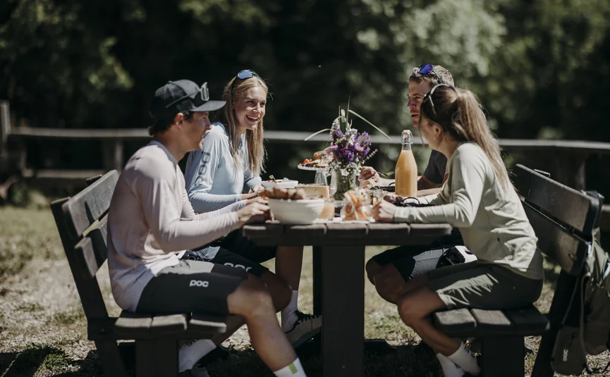 Vier personen zitten buiten aan een houten tafel en eten in de zon