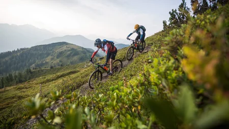 Was steckt hinter dem Umweltengagement von Bikehotels? Zwei Mountainbiker fahren auf einem schmalen Bergpfad bei bewölktem Himmel