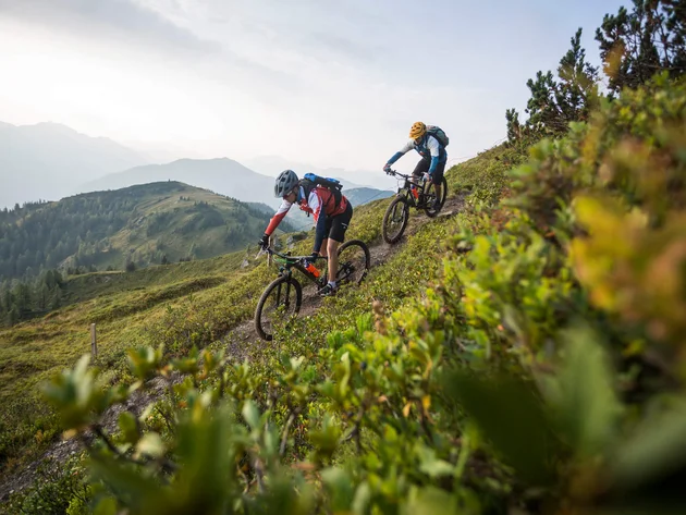 Was steckt hinter dem Umweltengagement von Bikehotels? Zwei Mountainbiker fahren auf einem schmalen Bergpfad bei bewölktem Himmel