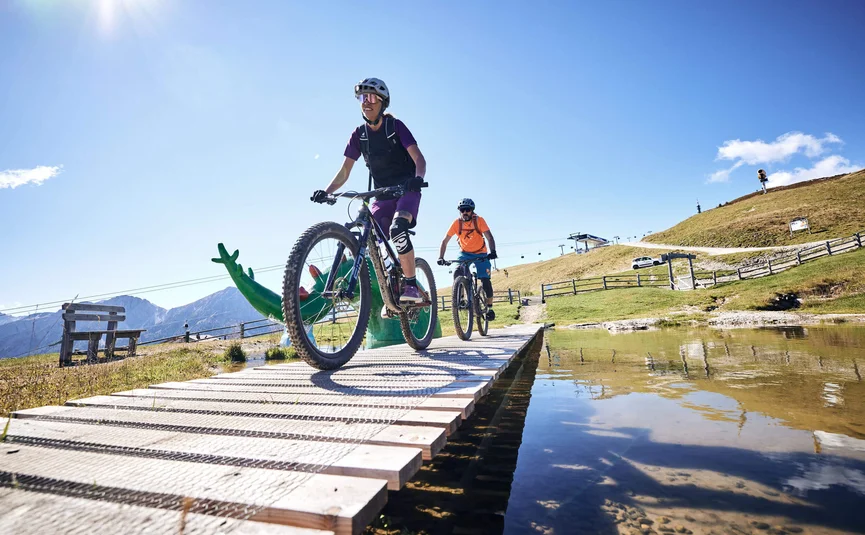 Zwei Radfahrer auf Holzsteg über Wasser bei sonnigem Wetter in den Bergen