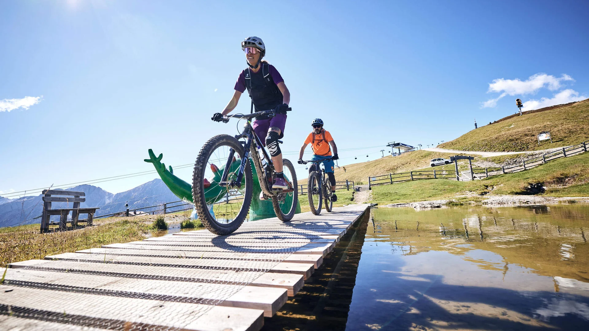 Zwei Radfahrer auf Holzsteg über Wasser bei sonnigem Wetter in den Bergen