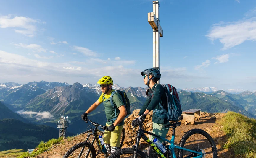 Twee mountainbikers met helmen bij een bergtopkruis in de Alpen