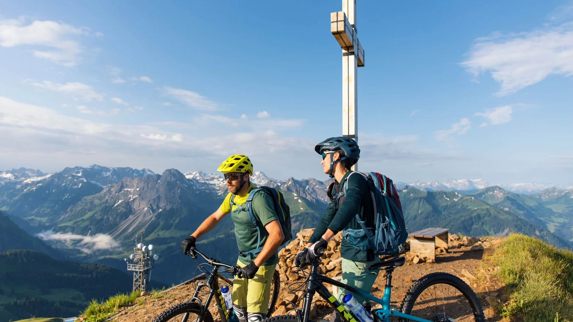 Zwei Mountainbiker mit Helmen an Berggipfelkreuz in Alpen