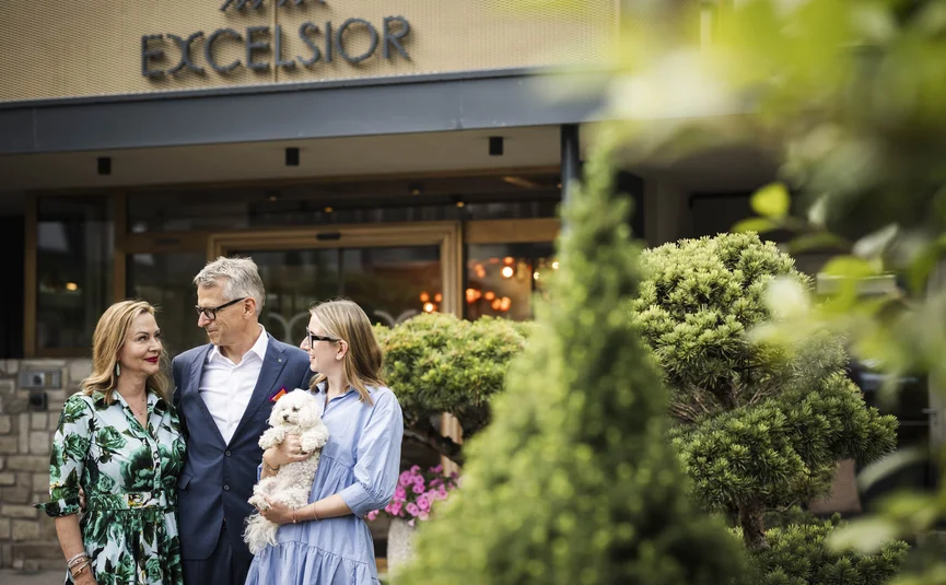 Three people with small white dog outside Excelsior building