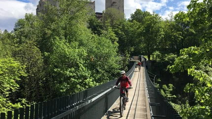 Cyclists crossing a suspension bridge with a castle in a lush green forest