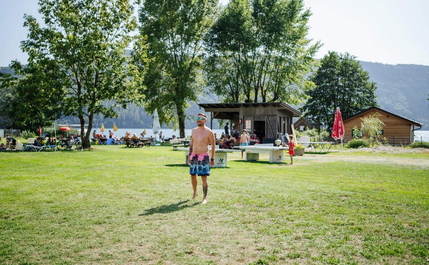 Man in zwembroek staat op grasveld bij meer met mensen en bomen