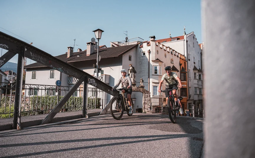 Zwei Radfahrer fahren auf einer Brücke in einer Altstadt bei sonnigem Wetter