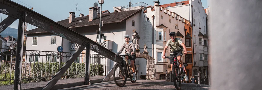 Zwei Radfahrer fahren auf einer Brücke in einer Altstadt bei sonnigem Wetter