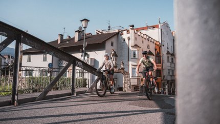 Bressanone © Brixen Tourismus - Hannes Engl Two cyclists riding on a bridge in a historic town on a sunny day