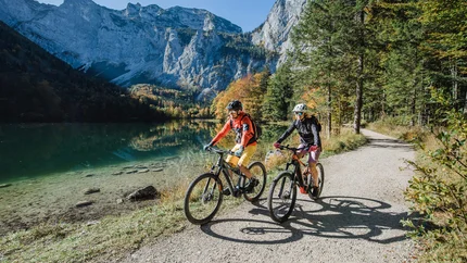 Twee fietsers op bospad naast bergmeer en bergen bij helder weer