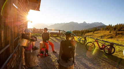 Drei Mountainbiker machen Pause bei einer Holzhütte mit Bergblick im Sonnenuntergang