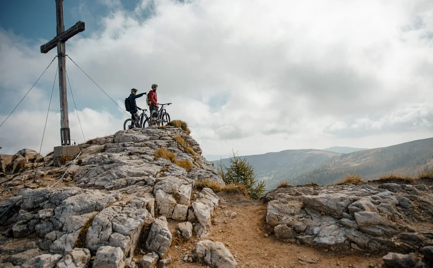 Two mountain bikers on rocky summit next to cross in the mountains