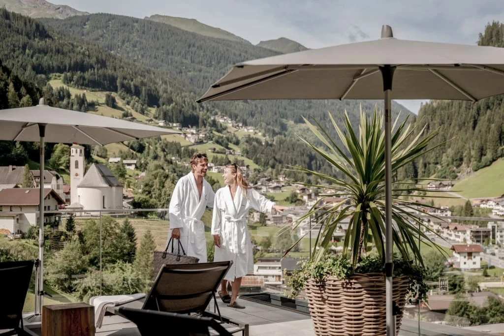 Zwei Personen im Bademantel auf einer Terrasse mit Sonnenschirmen und Bergblick