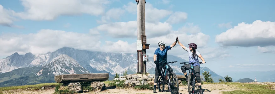 Zwei Mountainbiker geben sich bei einem Gipfelkreuz in den Bergen ein Highfive