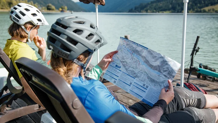Zwei Radfahrer lesen eine Karte am See mit Bergen im Hintergrund.