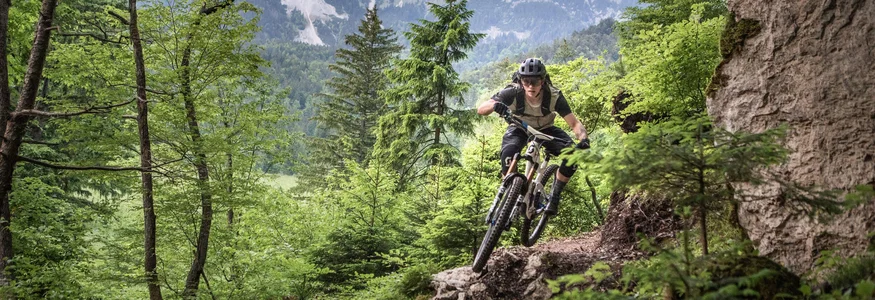 Dein Julische Alpen-Guide © Žirovnica - Jošt Gantar Mountainbiker fährt auf felsigem Waldpfad mit Bergkulisse