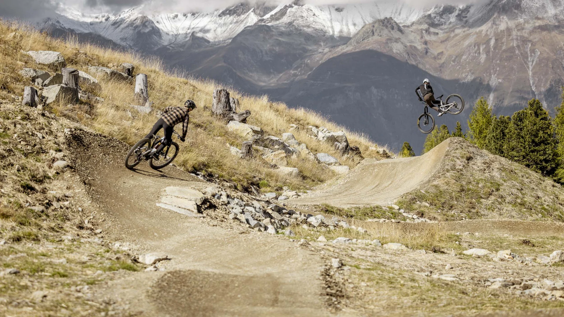 Zwei Mountainbiker fahren auf einem Bergtrail mit schneebedeckten Gipfeln im Hintergrund