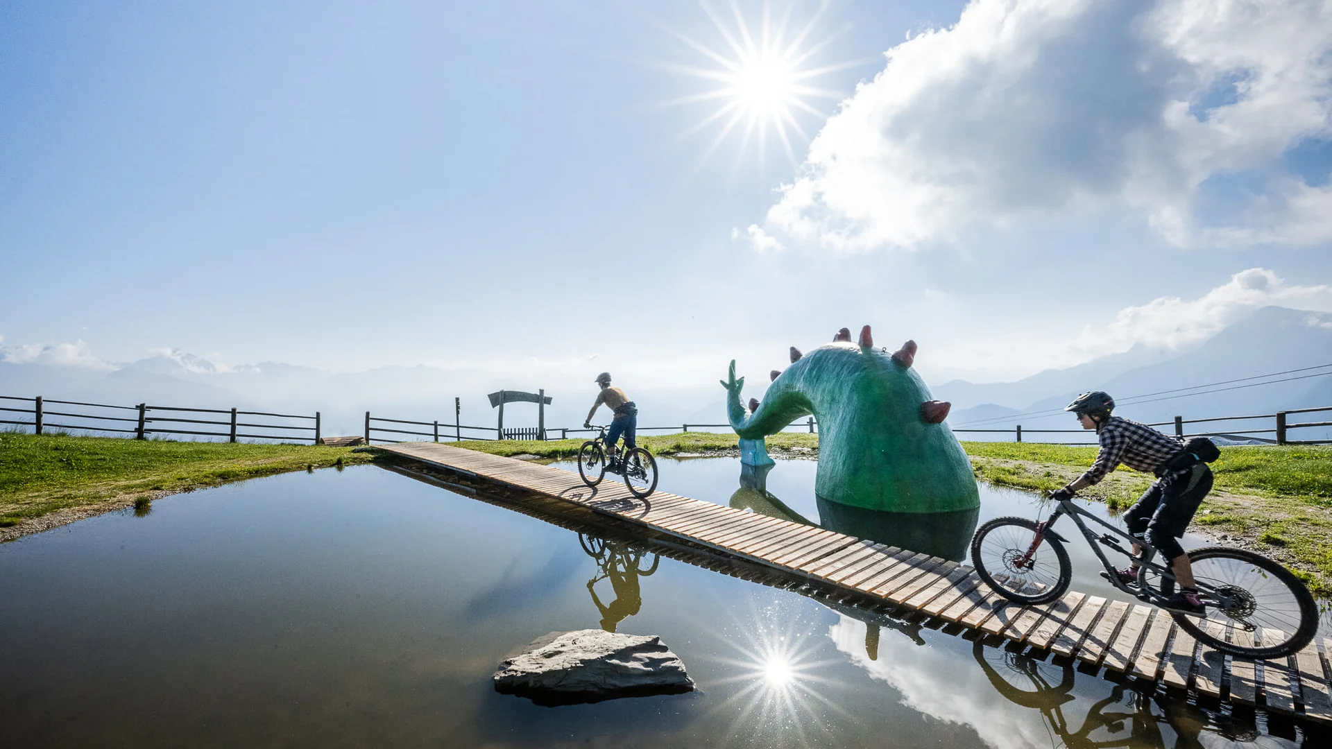 Zwei Radfahrer auf Brücke über Teich mit grüner Monster-Skulptur bei Sonne
