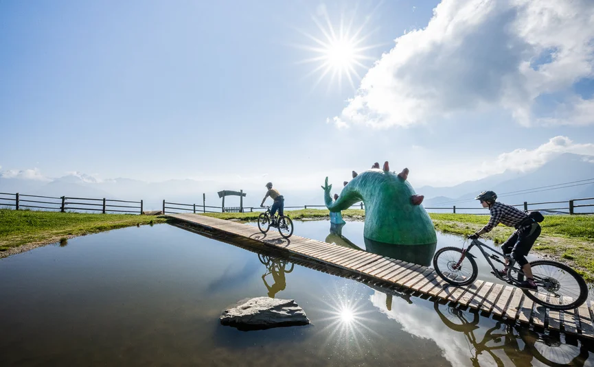Zwei Radfahrer auf Brücke über Teich mit grüner Monster-Skulptur bei Sonne