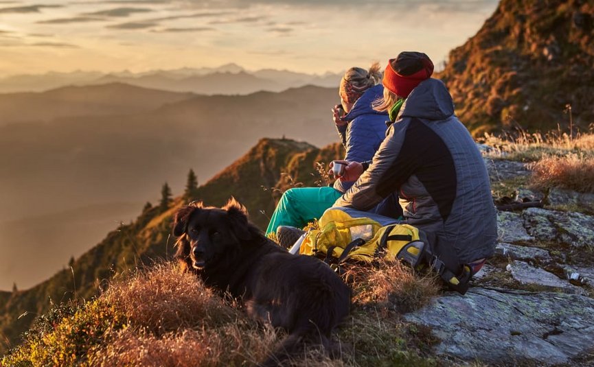 Twee mensen en een hond zitten op een berg bij zonsondergang