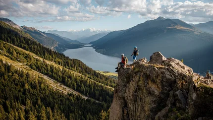 Zwei Wanderer auf Felsen mit Berg- und Seelandschaft im Hintergrund