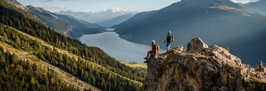 Zwei Wanderer auf Felsen mit Berg- und Seelandschaft im Hintergrund