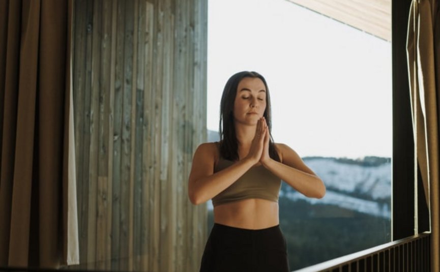 Frau meditiert mit geschlossenen Augen vor großem Fenster mit Bergblick