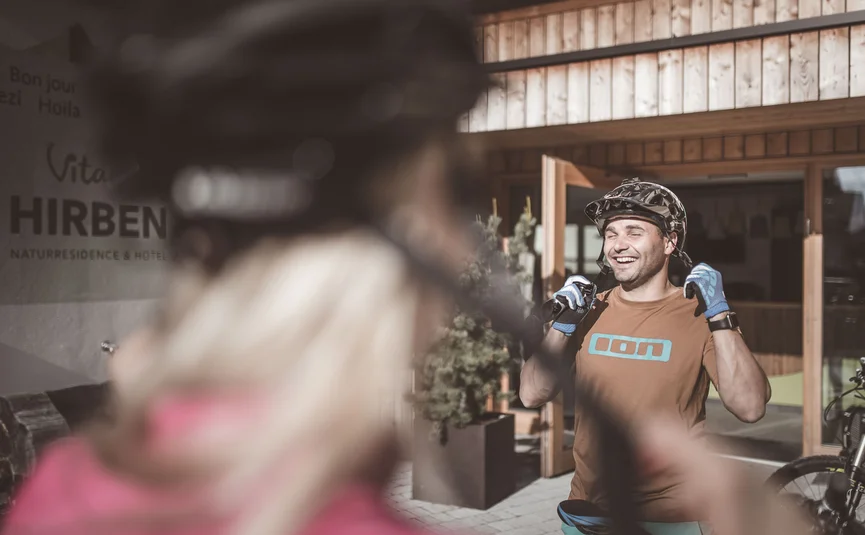 HIRBEN Naturlaub **** © Manuel Kottersteger Man wearing bike helmet and gloves smiling in front of hotel with wooden entrance