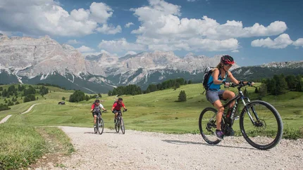 Three cyclists on gravel path with mountain backdrop on a sunny day