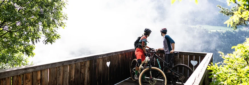 Two mountain bikers on a wooden viewpoint overlooking a foggy valley