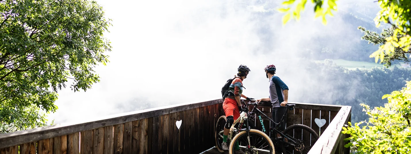 Two mountain bikers on a wooden viewpoint overlooking a foggy valley